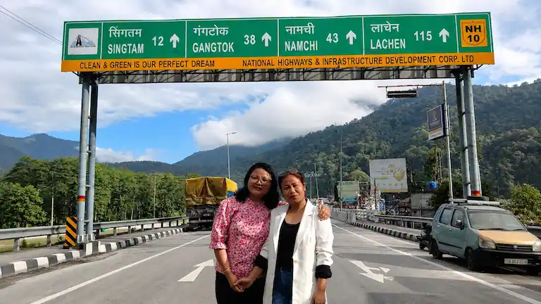 Travelers standing under the NH10 highway signboard showing distances to Gangtok and Namchi.